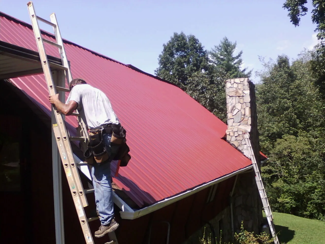 Roofer on a ladder beside a red metal roof and stone chimney — Soffit & Fascia Repair by Roofing Brothers in Senecaville